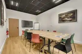 Salle de réunion moderne : grande table bois, chaises colorées, écran plat TV, tableau blanc et plafond acoustique noir.